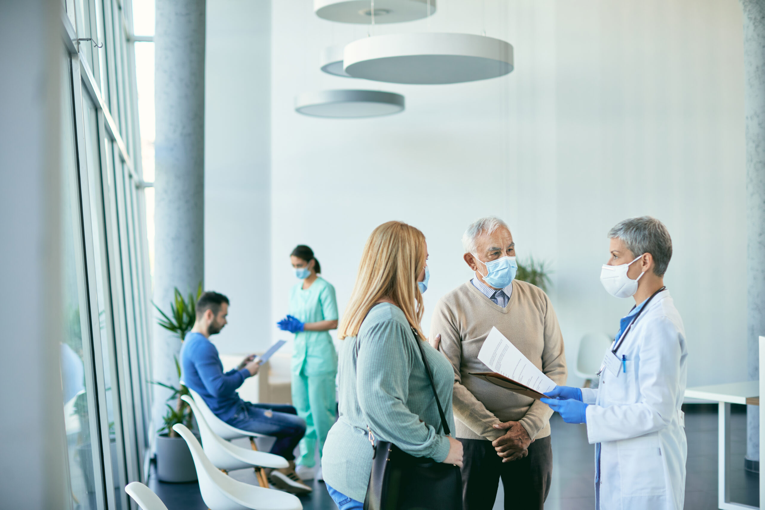 Female doctor and her patients wearing protective face masks while communicating about medical reports in hallway at the hospital. Focus is on senior man.