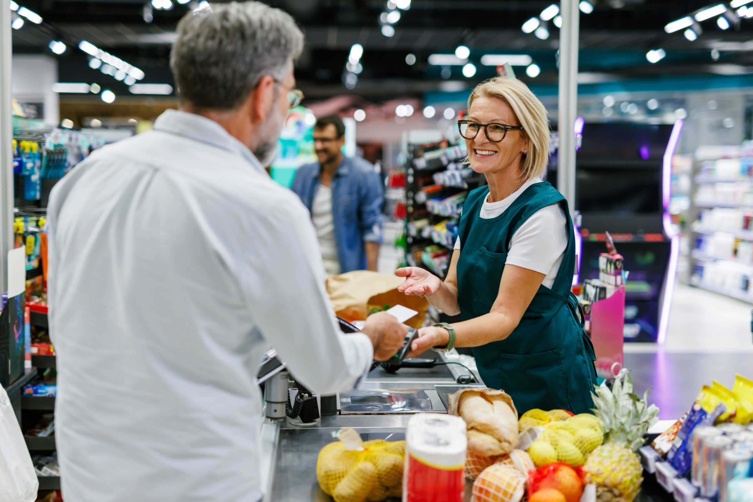 Senior customer paying with nfc technology on smartphone at supermarket checkout, while friendly smiling female cashier is holding pos terminal