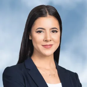 Eine Frau mit glattem dunklem Haar, die einen marineblauen Blazer über einem weißen Oberteil trägt, lächelt sanft vor einem hellblauen, verschwommenen Hintergrund - perfekt für Ihre nächste Kontakt ETI-Park-Einführung.