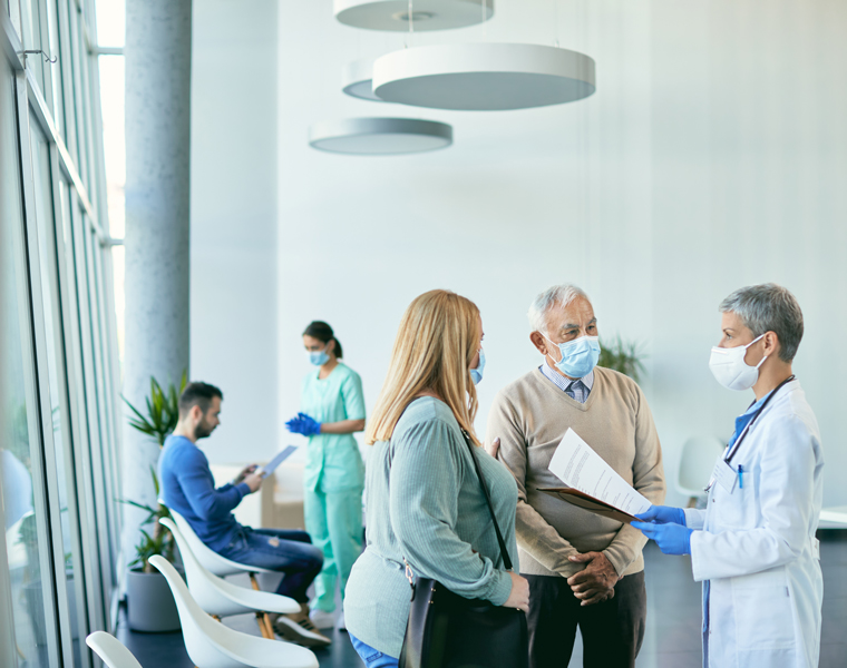 Ein Arzt mit Maske und Handschuhen spricht mit einem älteren Mann und einer Frau in einem modernen Wartebereich, während zwei weitere Personen, darunter eine Krankenschwester, im Hintergrund zu sehen sind. Alle tragen Masken.