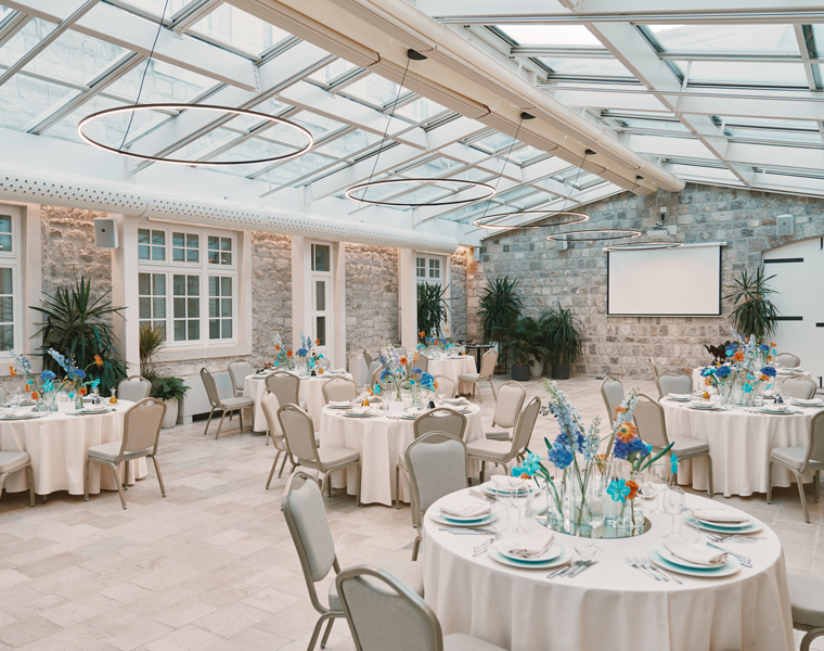 Ein heller, eleganter Veranstaltungsraum mit einer Glasdecke, runden Tischen für eine formelle Veranstaltung, weißen Tischdecken, hellen Stühlen, blauen und orangefarbenen Blumenkränzen und großen Fenstern entlang einer Steinwand.