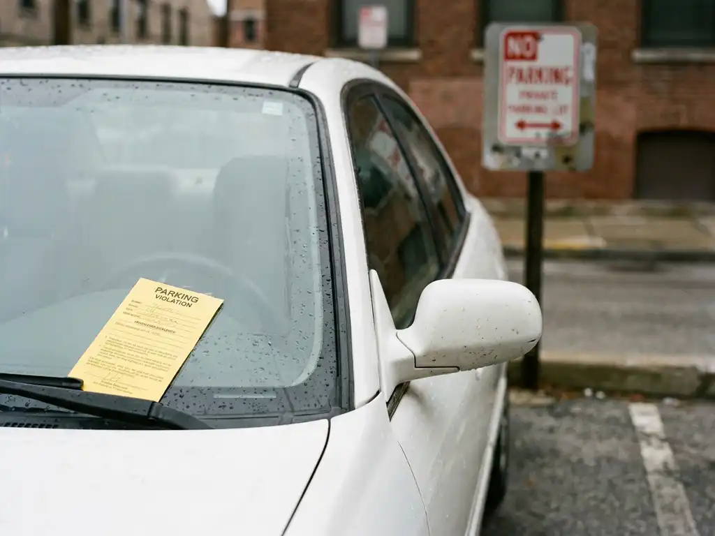 Parkverbot-Knöllchen unter einem Scheibenwischer auf privatem Parkplatz, Verbotsschild unscharf im Hintergrund.