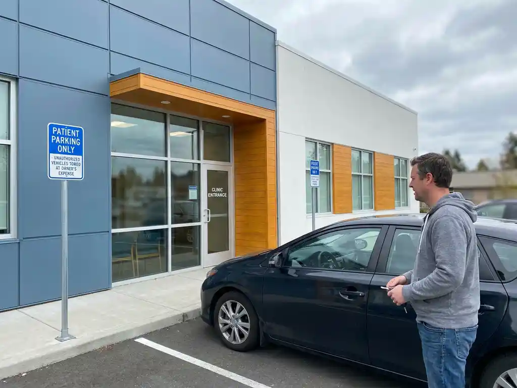 Frustrierter Patient mit Autoschlüssel neben besetztem Patientenparkplatz vor moderner Arztpraxis.